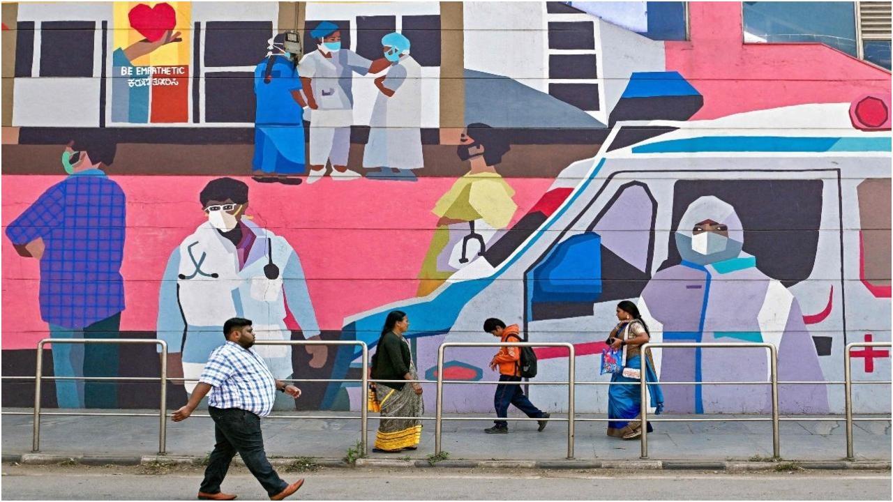 सोमवार को बेंगलुरु में फेस मास्क के इस्तेमाल के बारे में जागरूकता फैलाने वाली दीवार पर लगे भित्तिचित्र के पास से पैदल चलते लोग. तस्वीर/एएफपी